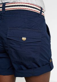 Pantalones cortos azul marino con dobladillo plegado, con una cintura tejida con rayas en rojo, blanco y azul. Incluyen un bolsillo trasero con un detalle de botón dorado.