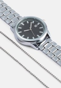 Sidabrinis nerūdijančio plieno laikrodis su juodu ciferblatu, švytinčiais valandų žymekliais ir datos rodymu. Šalia yra plona sidabrinė grandinėlė.