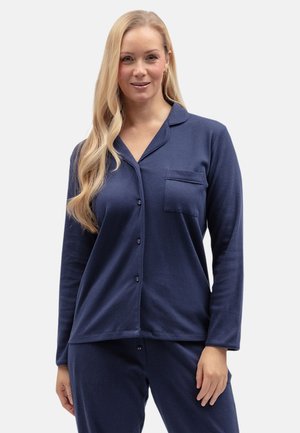 Frau mit langen blonden Haaren, die ein marineblaues, zum Knöpfen geeignetes Pyjamaoberteil und passende Hose trägt, steht vor einem schlichten hellen Hintergrund.