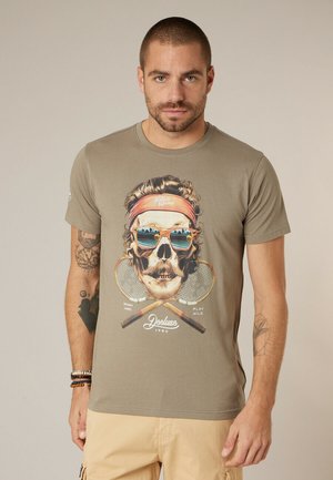 Man draagt een beige T-shirt met schedelafbeelding, zonnebril en hoofdband, gekruiste tennisrackets en lichte korte broek.