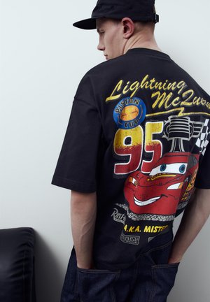 Ung mand iført sort kasket og sort T-shirt med stort, farverigt motiv af Lynet McQueen og nummer 95 fra filmen Cars.