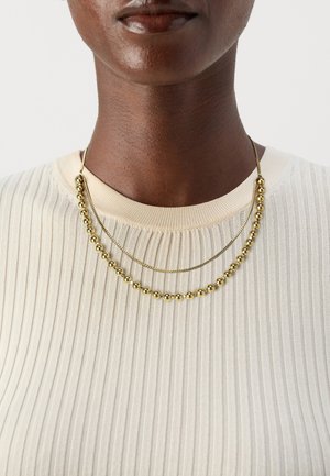 Collier en perles dorées à deux brins superposés, comprenant des perles rondes et une chaîne fine, présenté sur un haut léger à rayures.