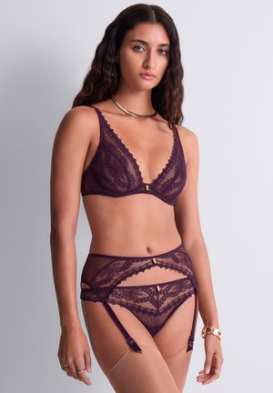 Jeune femme portant un ensemble de lingerie en dentelle violette avec porte-jarretelles et bas, debout devant un fond uni clair.