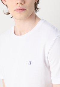 Tricou alb din bumbac cu guler rotund, având un mic logo brodat "II" în culori albastre pe partea stângă a pieptului. Textură netedă și mâneci scurte.