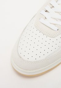 Hvit sneakers med perforert lær og semsket overdel, flate hvite snørebånd og såle i gummi. Rund tå og broderte detaljer.