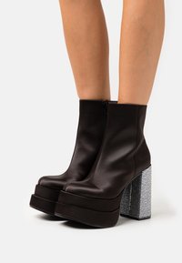 Botas de tobillo de plataforma negras con tacones gruesos cubiertos de rhinestones plateados, usadas sobre piernas desnudas.