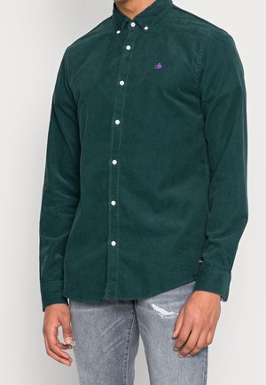 Mężczyzna w ciemnozielonej koszuli sztruksowej z małym fioletowym logo, w połączeniu z jasnoszarymi podartymi dżinsami, na jednolitym tle.