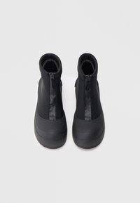 Pár černých voděodolných kotníkových bot s předními zipy a silnými gumovými podrážkami, pohled shora na světlém povrchu.