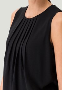 Schwarzes ärmelloses Oberteil mit vertikalen Falten, rundem Ausschnitt und glattem, leichtem Stoff. Keine sichtbaren Muster oder Beschläge.