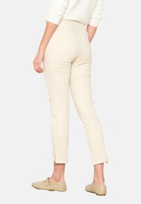 Beiga, åtsittande byxor med medelhög midja och blixtlåsdetalj vid vristerna. Tyget är slätt med en liten stretch. Två bakfickor synliga.