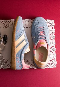 Baskets en suède bleu clair avec des accents roses et beiges, détails floraux décoratifs, posées sur une surface rouge avec un napperon en dentelle blanche en dessous.