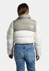 Giacca imbottita in beige chiaro e blocchi di colore grigio, con colletto alto e logo sulla schiena. Indossata con jeans a vita alta blu.