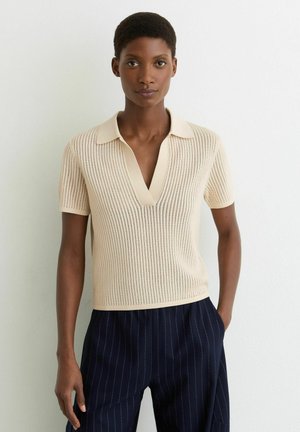 Vrouw met kort haar, gekleed in een beige gebreide polo met korte mouwen en marineblauwe broek met krijtstrepen, staand tegen een effen witte muur.