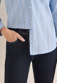 Lyseblå skjorte med vertikale striber og knapper, lige snit, kombineret med mørkeblå jeans med synlige sømme og forlommer.