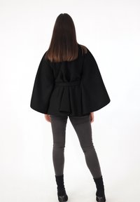 Schwarzer Mantel im Cape-Stil mit weiten Ärmeln, strukturiertem Stoff, tailliert mit Gürtel, getragen über grauer enger Hose und schwarzen Stiefeletten.