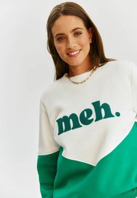 Bílá a zelená mikina se zaobleným designem. Na přední straně je vytlačený text "meh." v zelené barvě. Hladká látka s uvolněným střihem.