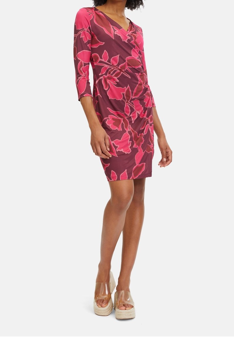 Femme portant une robe portefeuille fleurie violette et rose avec des manches trois-quarts et des sandales compensées transparentes, debout sur un fond blanc.