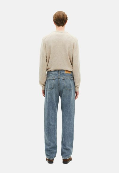 Suéter beige de manga larga combinado con jeans ligeros y sueltos que tienen un parche en la parte trasera. Un calzado oscuro completa el atuendo.