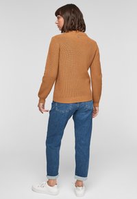 Koperkleurige gebreide trui met een detail achter op de kraag, gecombineerd met blauwe opgerolde jeans en witte sneakers. De stof heeft een gestructureerde afwerking.