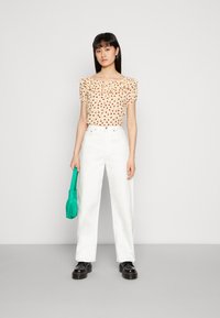 Blusa floral com decote franzido em creme, combinada com calças brancas de perna larga de cintura alta e sapatos de plataforma pretos. Segurando uma bolsa verde.