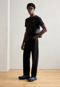 Černé tričko s krátkým rukávem, černé kalhoty a modro-černé boty. Model má sluneční brýle a malou černou přes rameno tašku. Jednoduchý, moderní design.