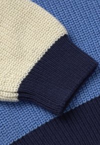 Gros plan sur un pull en tricot bleu avec une manche crème et un détail de poignet côtelé bleu foncé.