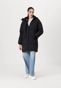 Black padded long coat with a hood, featuring a zip front, large pockets, and a quilted texture, worn over light blue jeans and white shoes.