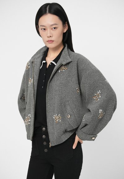 Jauna sieviete, tērpta pelēkā jaku ar ziedu spīdošiem akmentiņiem, melnā pogātā krekliņā un melnās augstās bikšu bikses, stāv ar roku kabatā.