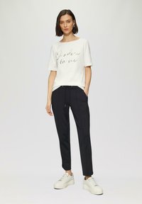 Witte t-shirt met korte mouwen en een gouden opdruk met de tekst "J'adore la vie." Gecombineerd met zwarte tapered broek en witte platform sneakers.