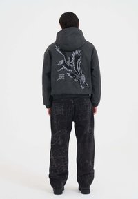 Persona che indossa una felpa nera corta con una grande grafica di aquila e pantaloni neri larghi con sottili stampe di serpente e ragnatela, in piedi girato di spalle.