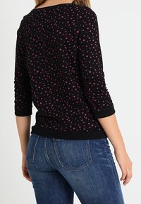 Top lungo nero a maniche lunghe con piccoli motivi floreali rosa, scollatura rotonda, polsini e vita a coste. Abbinato a jeans blu.