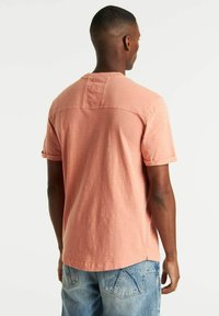 T-shirt a maniche corte, di un leggero rosa, con scollo rotondo e risvolti, caratterizzato da un pannello posteriore strutturato e una vestibilità comoda. Abbinato a pantaloni di jeans corti.