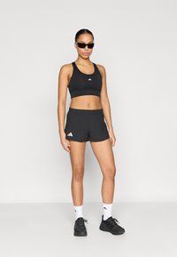 Tenue de sport noire comprenant un crop top ajusté et un short ample, tous deux ornés d'accents de logo blancs. Des baskets noires et des chaussettes blanches complètent le look.