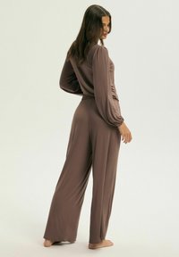 Brązowy, długi kombinezon z talią ściągniętą i szerokimi nogawkami. Miękka, gładka tkanina o delikatnym połysku. Brak widocznych wzorów lub elementów metalowych.