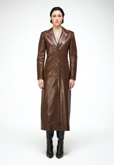 Nő áll egy hosszú barna bőrkabátban, amelynek hajtókája van, elöl gombos, bézs színű belső felső ruhadarabbal, valamint fekete térdig érő csizmában, fehér háttér előtt.
