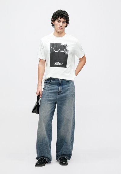 Biele tričko s čiernym grafickým potlačou a textom „Milano“. Nosené s svetlomodrými džínsami s rozšírenými nohavicami. Model drží čiernu tašku, na nohách má čierne topánky.