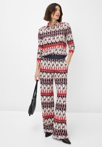 Completo in crema, rosso e navy composto da blusa a fantasia e pantaloni. La blusa ha un colletto; i pantaloni sono svasati con una cintura nera. Accessorio: borsa nera.