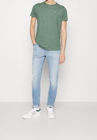 Mężczyzna w zielonej koszulce z krótkim rękawem, jasnoniebieskich dżinsach z mankietami, białych sneakersach New Balance, z jedną ręką w kieszeni, stoi na szarym podłożu.