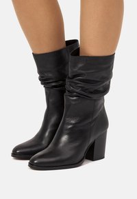 Botines de cuero negro con un diseño holgado y punta puntiaguda. Tacón cuadrado que aporta estabilidad. Textura suave y detalles mínimos.