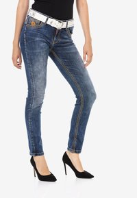 Schmale blaue Denim-Jeans mit Ausbleichungen, gelben Nähten und einem weißen mit Nieten versehenen Gürtel. Getragen mit schwarzen Stiletto-Absätzen.