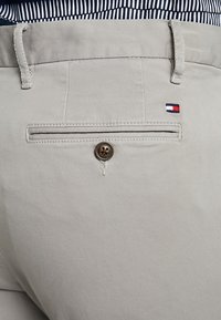 Světle šedé kalhoty s zadní kapsou na knoflík a malou cedulkou s logem v červené, bílé a modré barvě. Hladká textura látky.