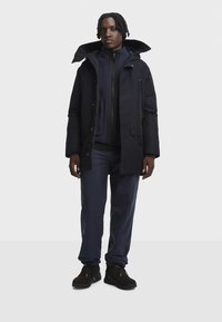 Tumesinine parka suurte kapuutsiga, ees tõmblukuga ja mitmete taskutega, kantud sobiva tõmblukuga jaku üle, sellega koos tumesiniste pükste ja tumedate kingadega.