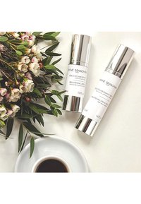 Two glass cosmetic bottles with silver caps: a brightening serum and cream, labeled in black text, beside green leaves and white flowers.