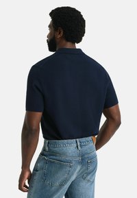 Marinblå kortärmad polotröja med en strukturerad mönstring. Bärs tillsammans med ljusblå jeans med bakfickor och en detaljerad läderbälte.