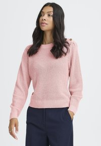 Femme avec de longs cheveux noirs portant un pull en tricot rose avec des détails de boutons, un pantalon marine et une bague à la main gauche, sur un fond uni.
