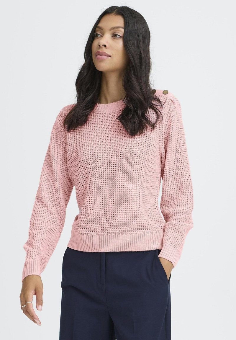 Femme avec de longs cheveux noirs portant un pull en tricot rose avec des détails de boutons, un pantalon marine et une bague à la main gauche, sur un fond uni.