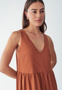 Vestido sin mangas en color óxido con escote en V profundo, falda acampanada con detalle fruncido en la cintura y una superficie de tejido texturizado.