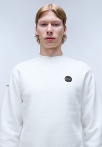 Felpa bianca a girocollo realizzata in tessuto morbido. Presenta una patch circolare con logo sul petto e un design minimalista con collo a costine.