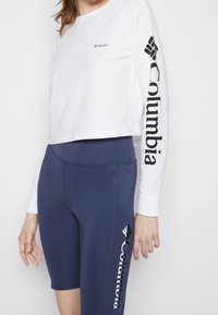 Personne portant un crop top blanc à manches longues et un short cycliste bleu marine taille haute, tous deux avec des logos Columbia noirs sur les manches et la jambe.