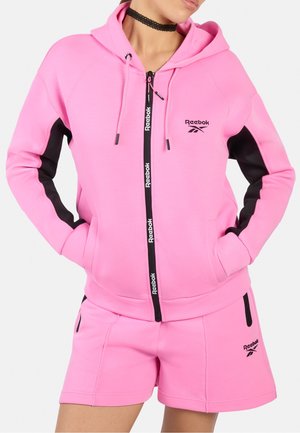 Femme portant un sweat à capuche zippé Reebok rose vif avec des accents noirs sur les côtés et un short rose assorti avec des poches zippées noires.